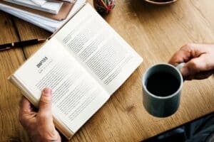 Person reading a book and holding a coffee mug on a wooden desk.