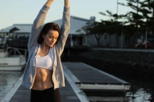 Modern woman stretching outdoors at sunset, near a marina with boats and houses in the background.